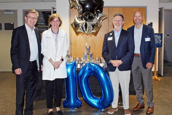 Four individuals stand together, with blue "1" and "0" balloons between them in celebration
