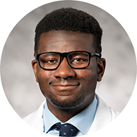 Dr. Kevin Ig-Izevbehai; black male, black-rimmed glasses, light-blue botton-down shirt, dark-blue tie with light dots, white doctor's coat.