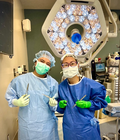 Two individuals in surgical attire stand together smiling inside an operating room