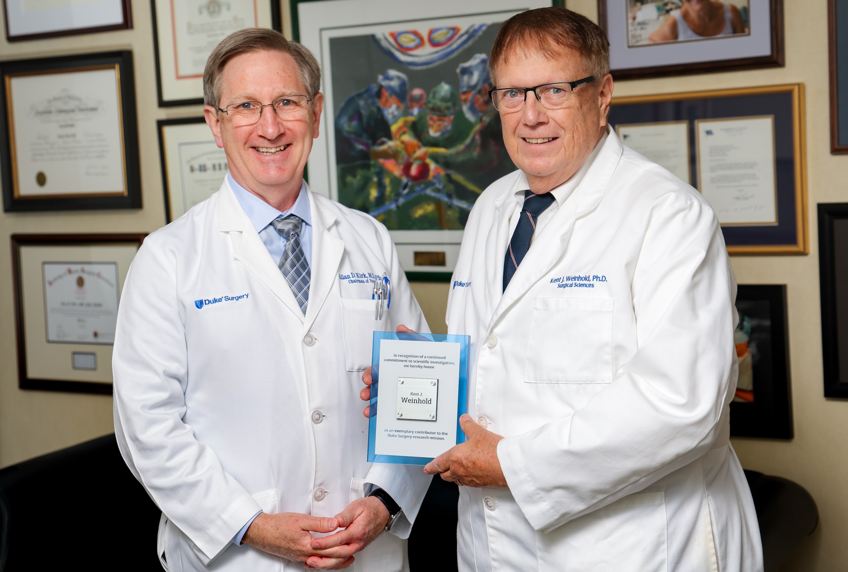 Dr. Allan Kirk and Dr. Kent Weinhold stand in their white coats smiling at the camera as Dr. Weinhold is presented with a plaque