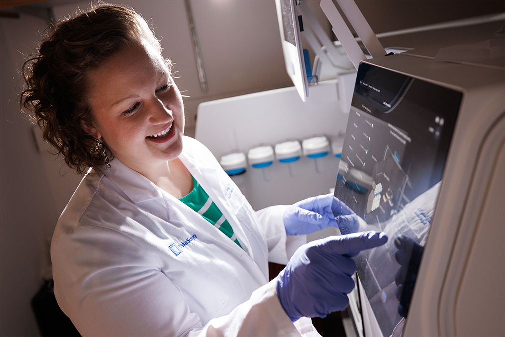 A woman in a white lab coat and blue gloves smiles as she works in her laboratory