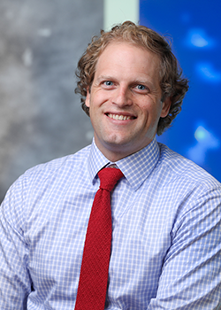 A man in a blue gingham button-down shirt and red tie smiles at the camera