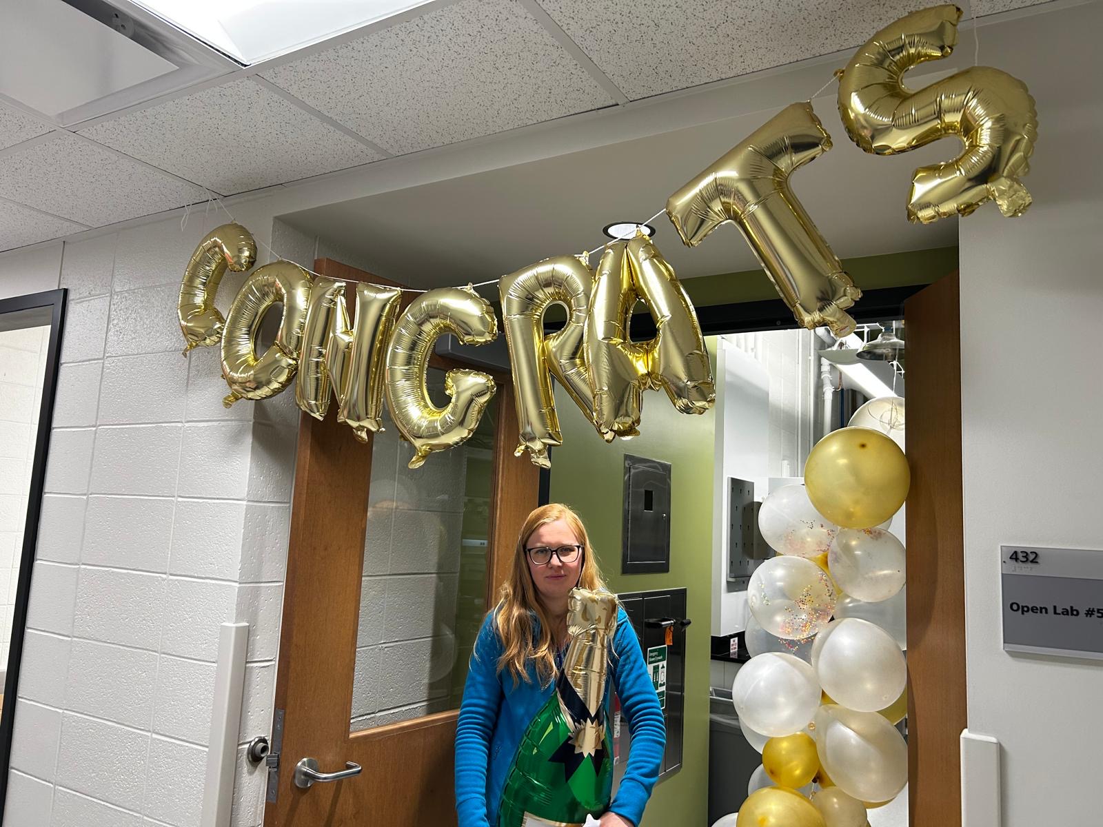 A woman stands smiling under balloons that spell out "Congrats"