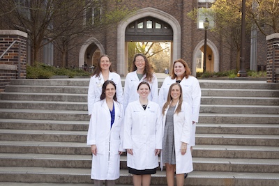 Six women in white coats stand on outdoor steps and pose for a photo