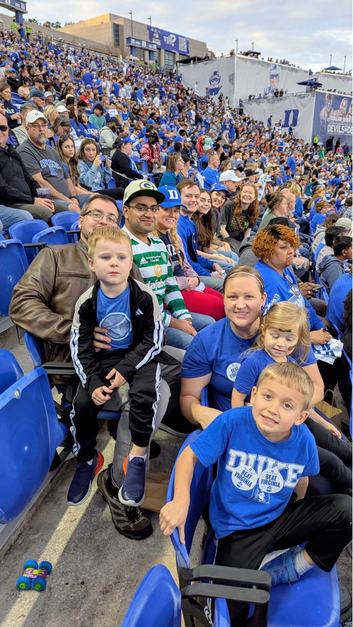 A group of people and their children pose for a photo at a Duke football game. They are all wearing Duke blue