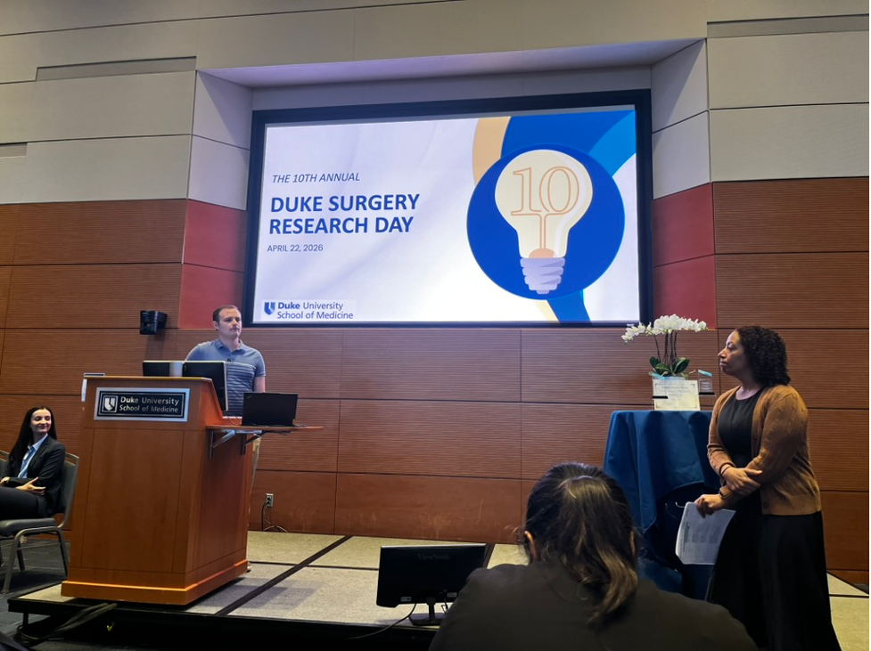 A man stands at a podium on stage giving a presentation. The image on the screen behind him reads "Duke Surgery Research Day"