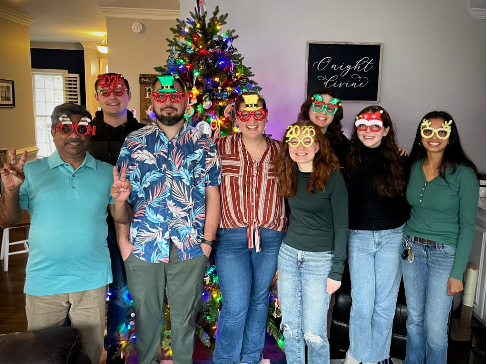 A group of 8 people pose together in front of a Christmas tree, and they are all wearing fun Christmas and holiday themed glasses
