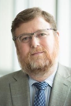 Headshot photo of Dr. Bruce Sullenger, wearing his glasses and a light grey suit and blue checked tie