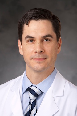 Dr. Jacob Schroder wears a white coat and poses for a professional headshot photo in front of a grey backdrop