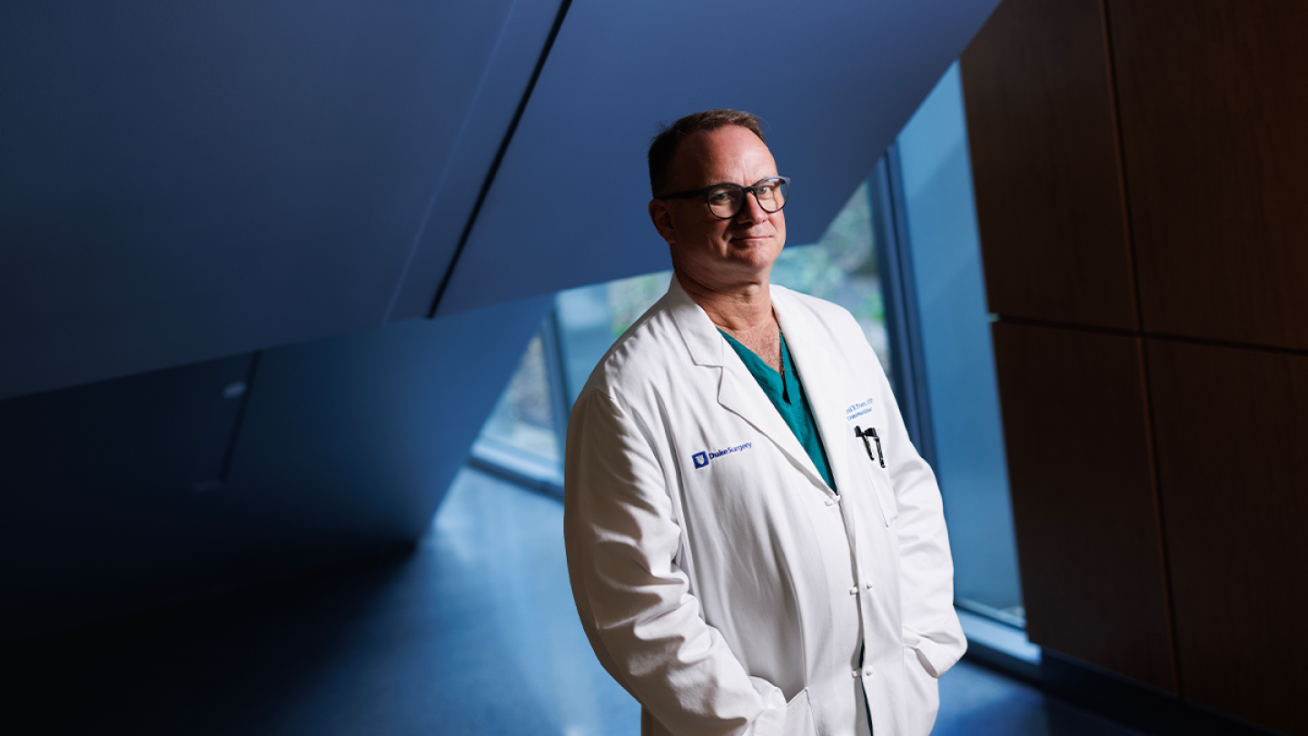 A man in a white physician coat stands in a hospital hallway