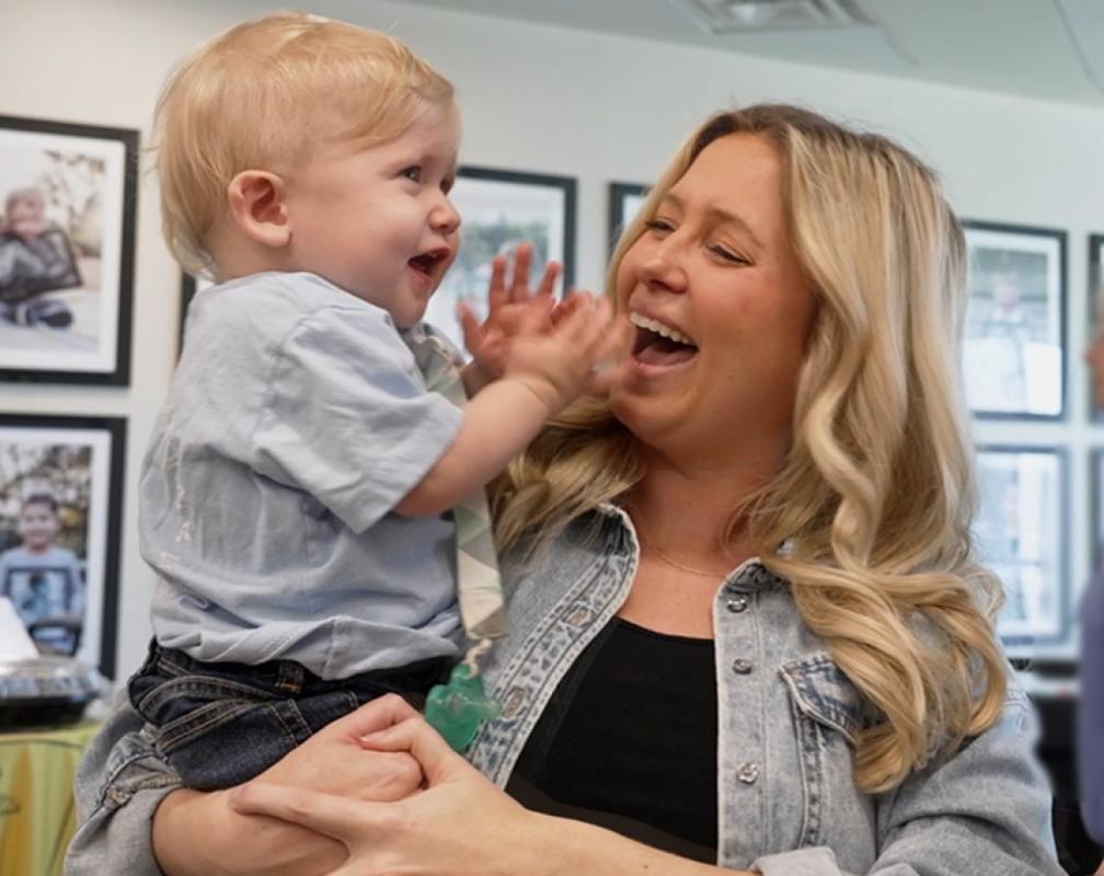 A smiling woman holds her young child, who is clapping his hands