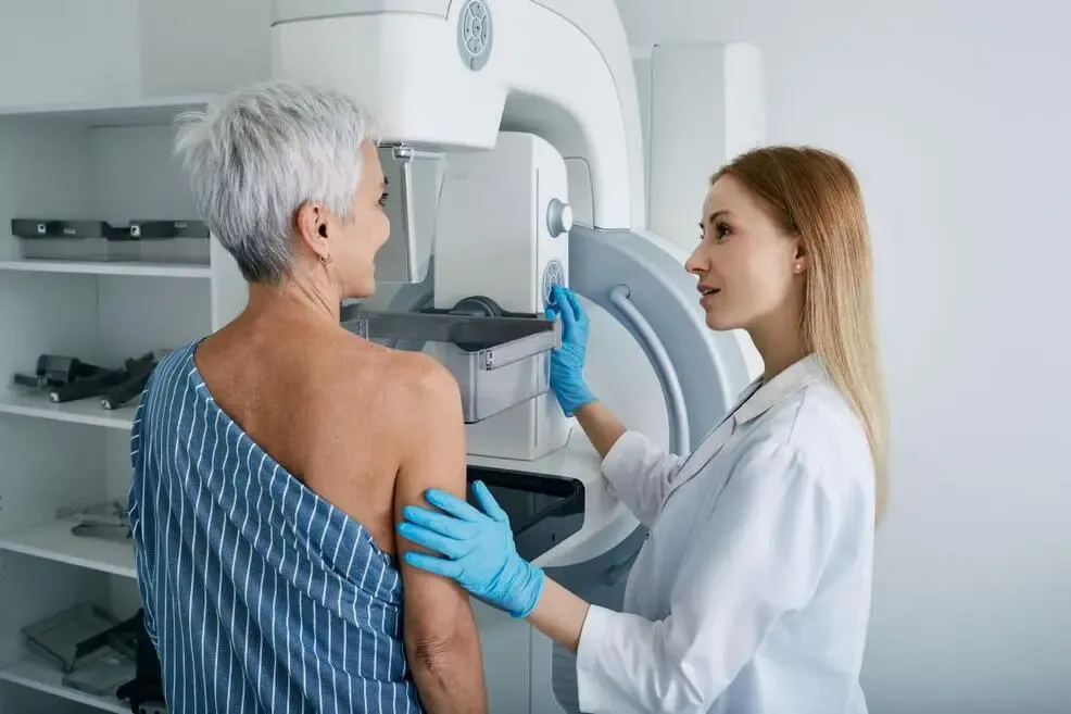 A blonde woman in a white coat prepares another woman for a mammogram screening