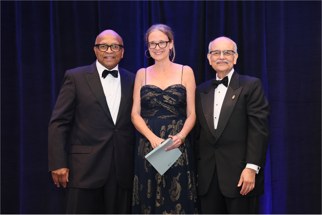 Two men and a woman in formal attire pose in front of a blue velvet backdrop at an awards ceremony