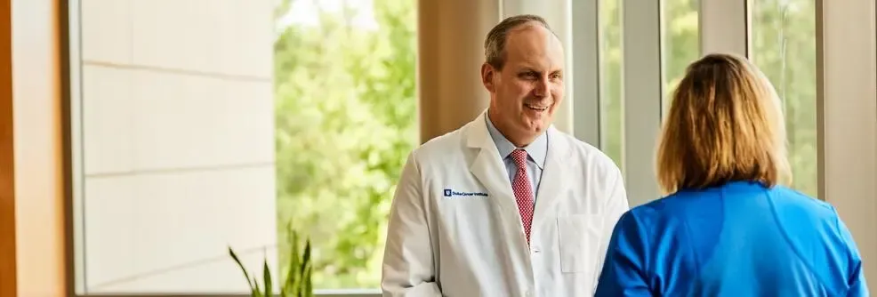 A male physician in a white coat smiles while speaking to a patient
