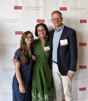 A woman poses between her husband and daughter in front of a backdrop that features the logo for the Boston University School of Medicine