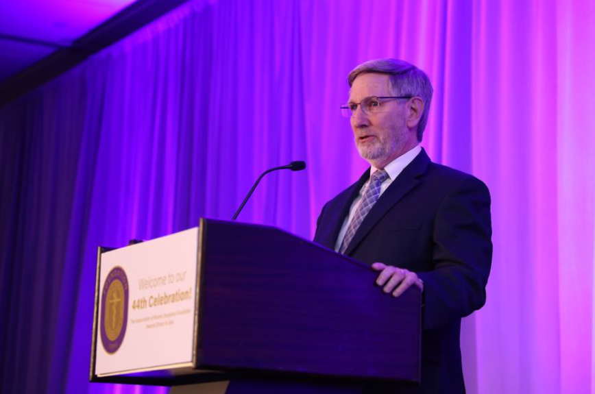 Dr. Allan Kirk stands at a podium at the Association of Women Surgeons conference, and the stage is lit by purple and pink lighting