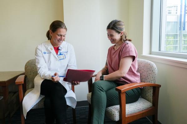 A female physician wears a white coat, while sitting in a chair discussing a chart with a female patient next to her