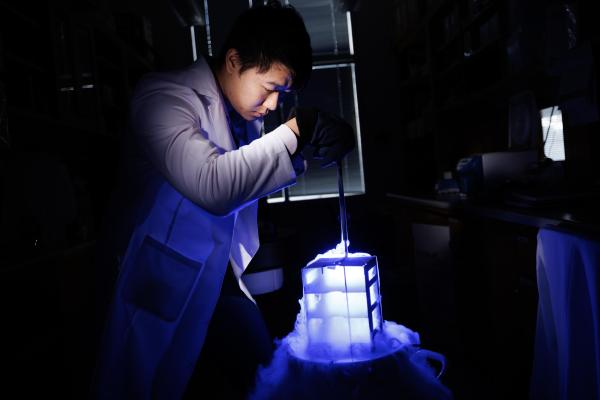 A technician works in a dark laboratory, over a device that emits a glowing blue light