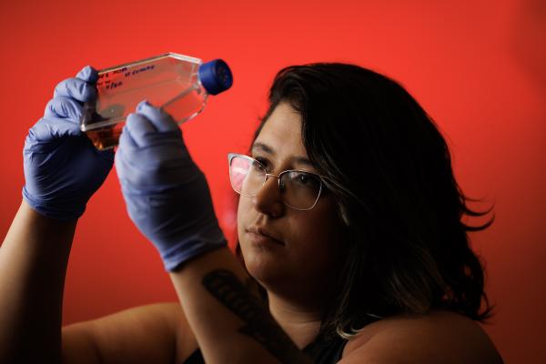 A woman with dark hair and wearing blue sterile gloves examines a bottle, against a red-lit background