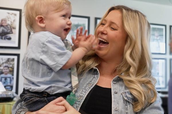 A smiling woman holds her young child, who is clapping his hands