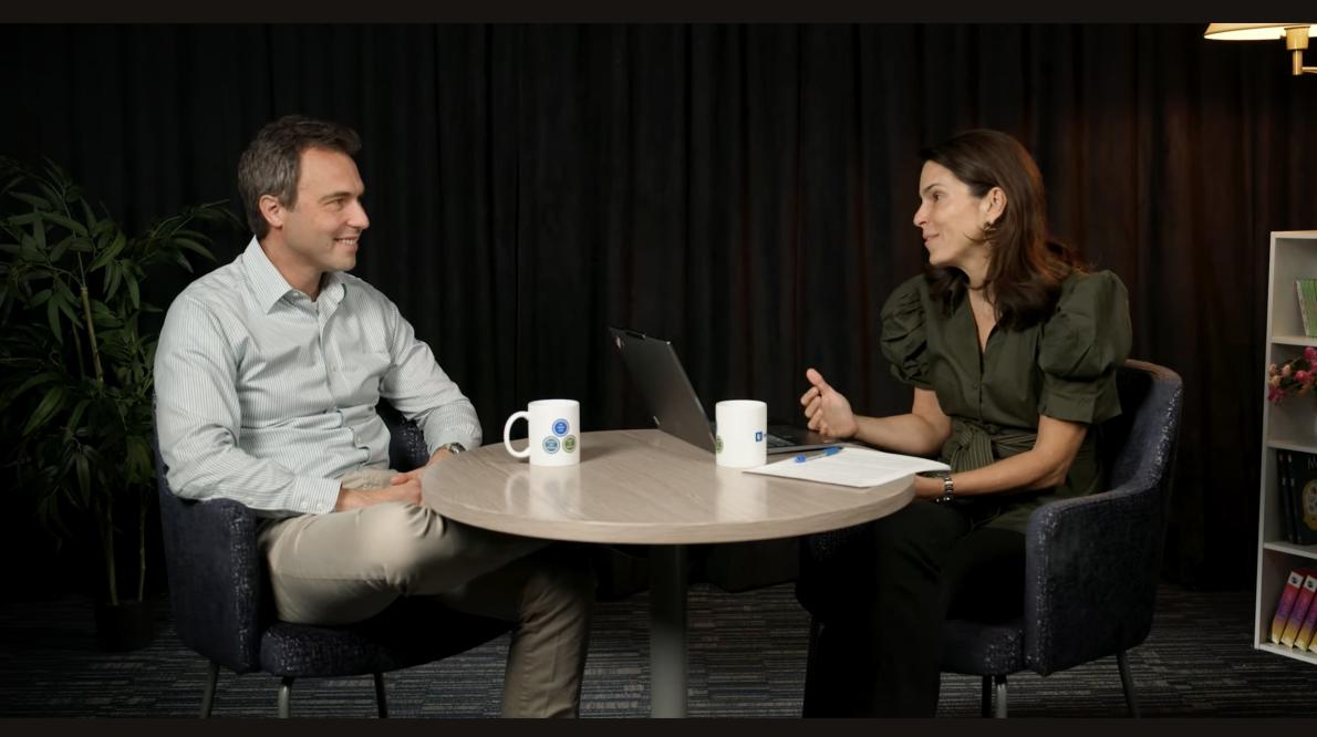 A man (left) and a woman (right) sit and talk at a round table in front of a black backdrop
