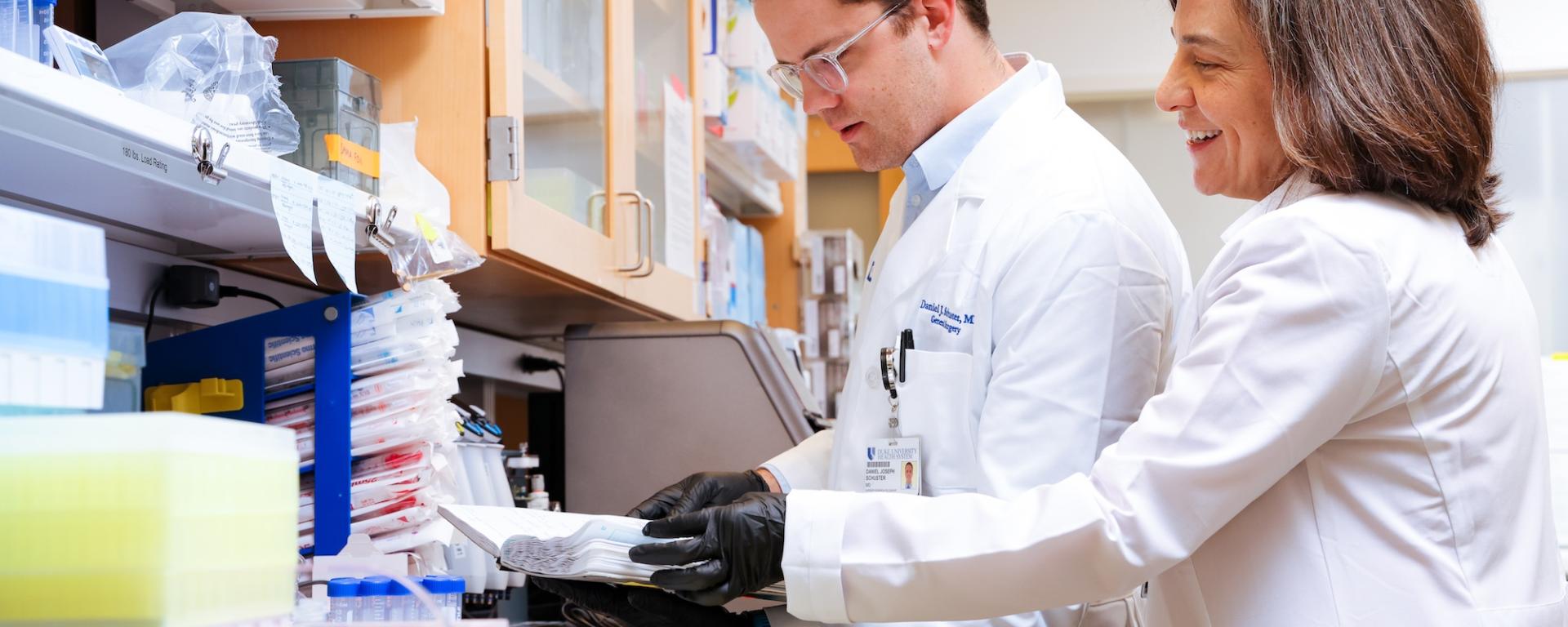 Dr. Georgia Tomaras (right) stands in a lab with Dr. Daniel Schuster (left) examining lab specimens
