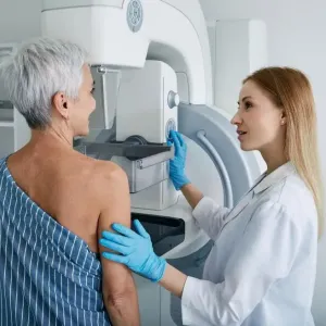 A blonde woman in a white coat prepares another woman for a mammogram screening